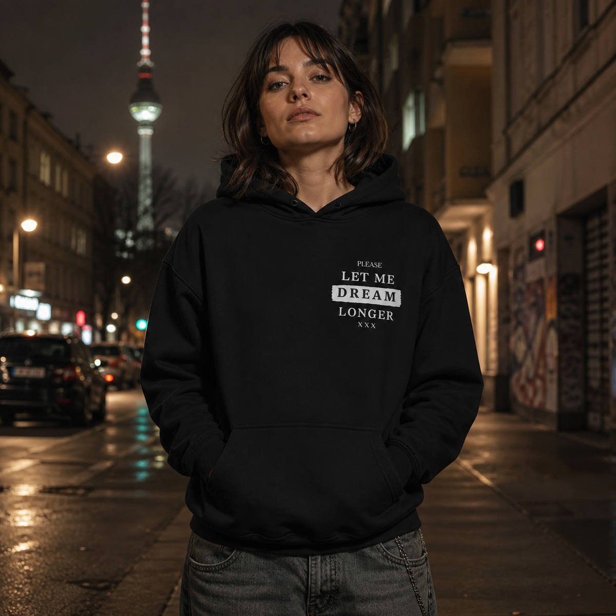 Weibliches Model trägt den schwarzen Dreamers Graphic Hoodie von NiZED mit Let Me Dream Longer Frontprint bei Nacht in Berlin – KI-generierte Darstellung